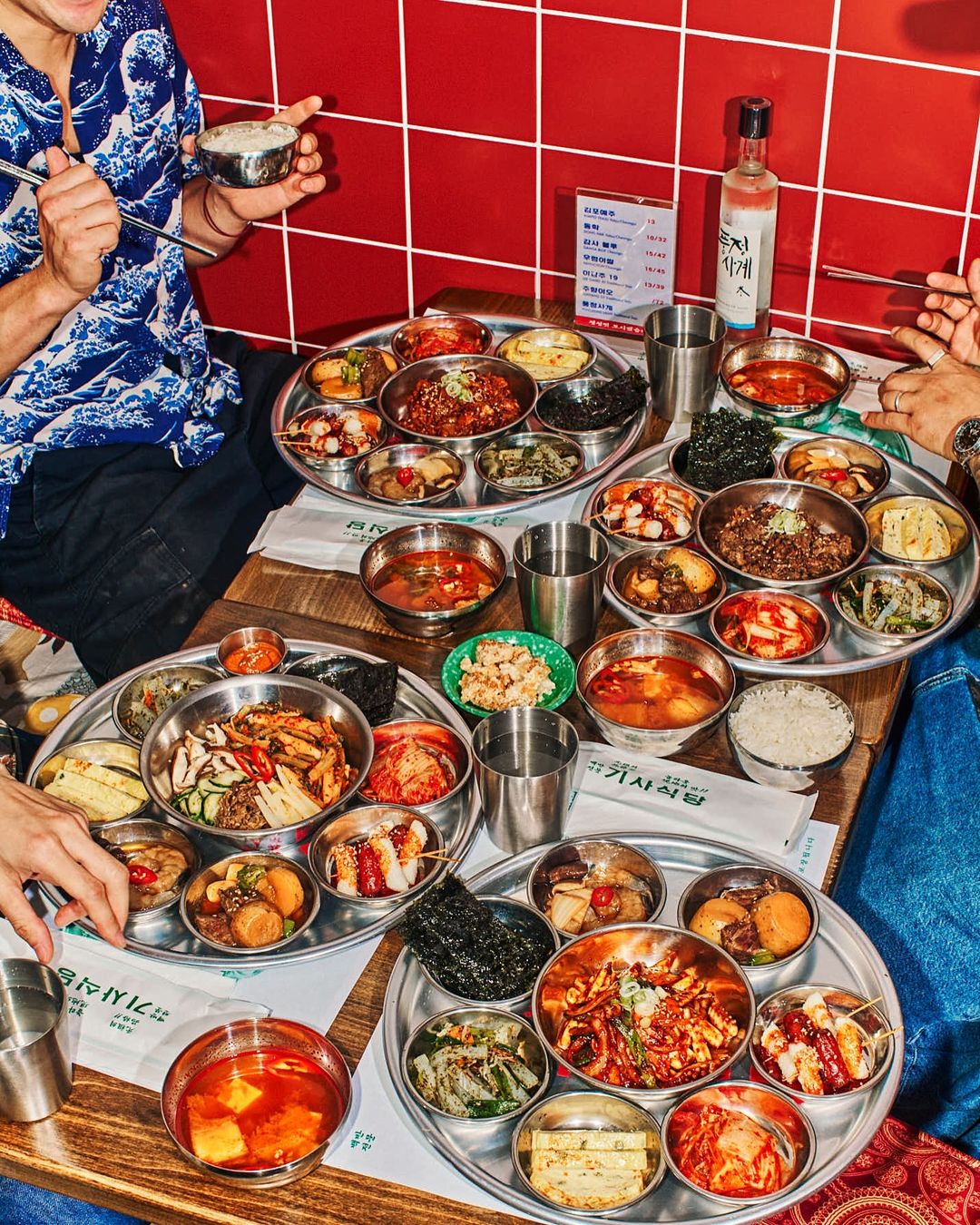 Overhead shot of group dining around a table filled with stainless steel trays and bowls of Korean side dishes with a red tiled background.