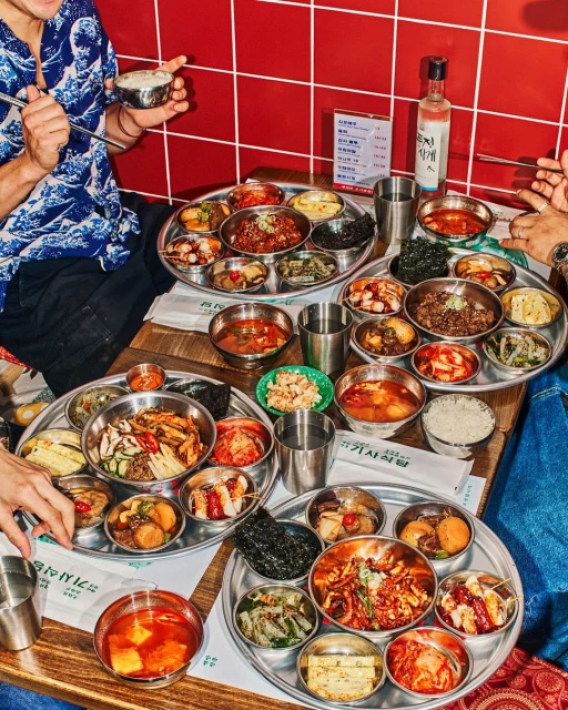 Overhead shot of group dining around a table filled with stainless steel trays and bowls of Korean side dishes with a red tiled background.