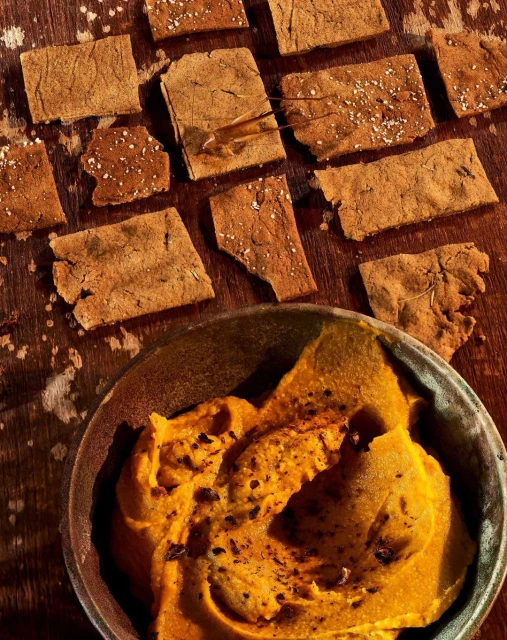 Overhead shot of rectangular crackers sprinkled with salt and a bowl of golden dip on a rustic wood surface, highlighting food photography.