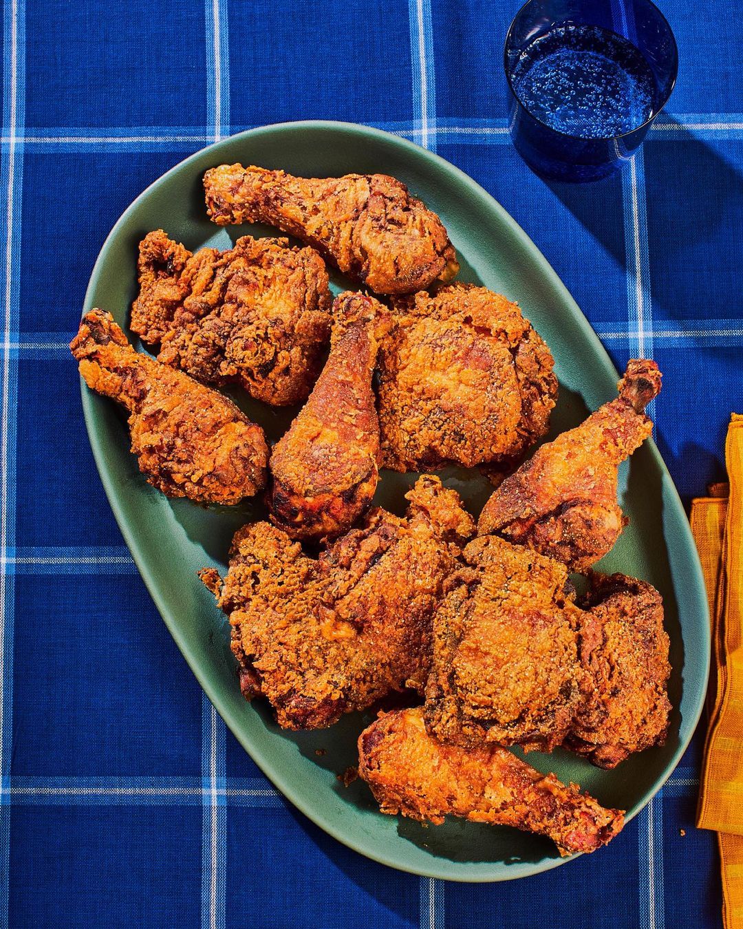 Overhead shot of fried chicken pieces on a green platter, set on a blue plaid tablecloth with a glass of water and yellow napkin.