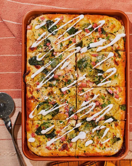 Overhead shot of a rectangular flatbread pizza cut into slices in a metal pan, featuring toppings such as spinach, zucchini, and a white sauce drizzle.