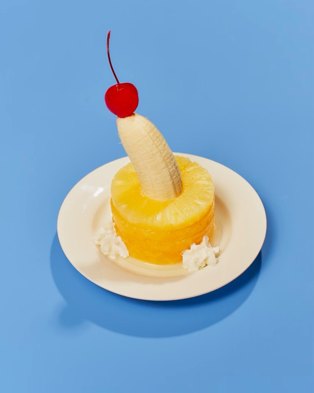 Festive dessert: Pineapple slice topped with banana and cherry, adorned with dollops of whipped cream on a white plate, blue background.