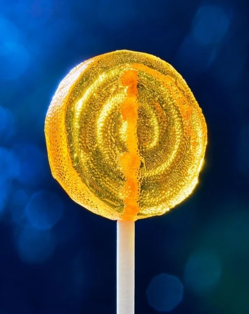 Close-up of a vibrant yellow lollipop with a clear, round swirl design, mounted on a white stick, set against a blurred blue background.