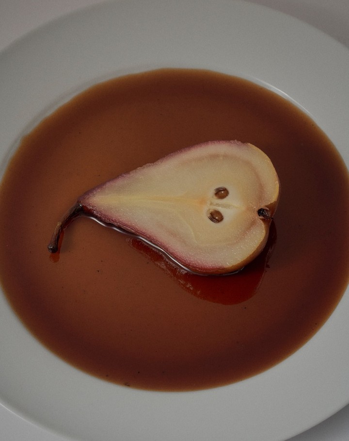 Close-up of a half-cut poached pear on a white plate, surrounded by brown sauce, capturing its unique form, seeds and texture.