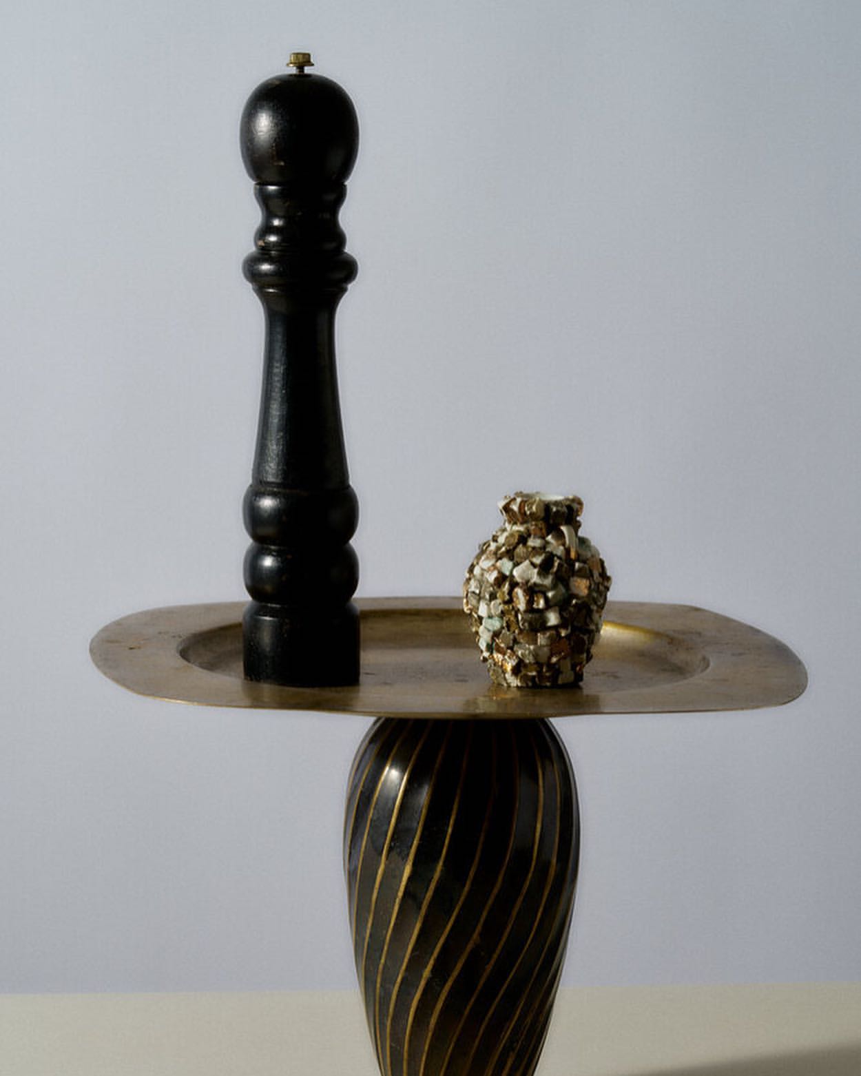 Still life featuring a black pepper mill, a textured vase, and a gold tray on a pedestal, set against a neutral background.