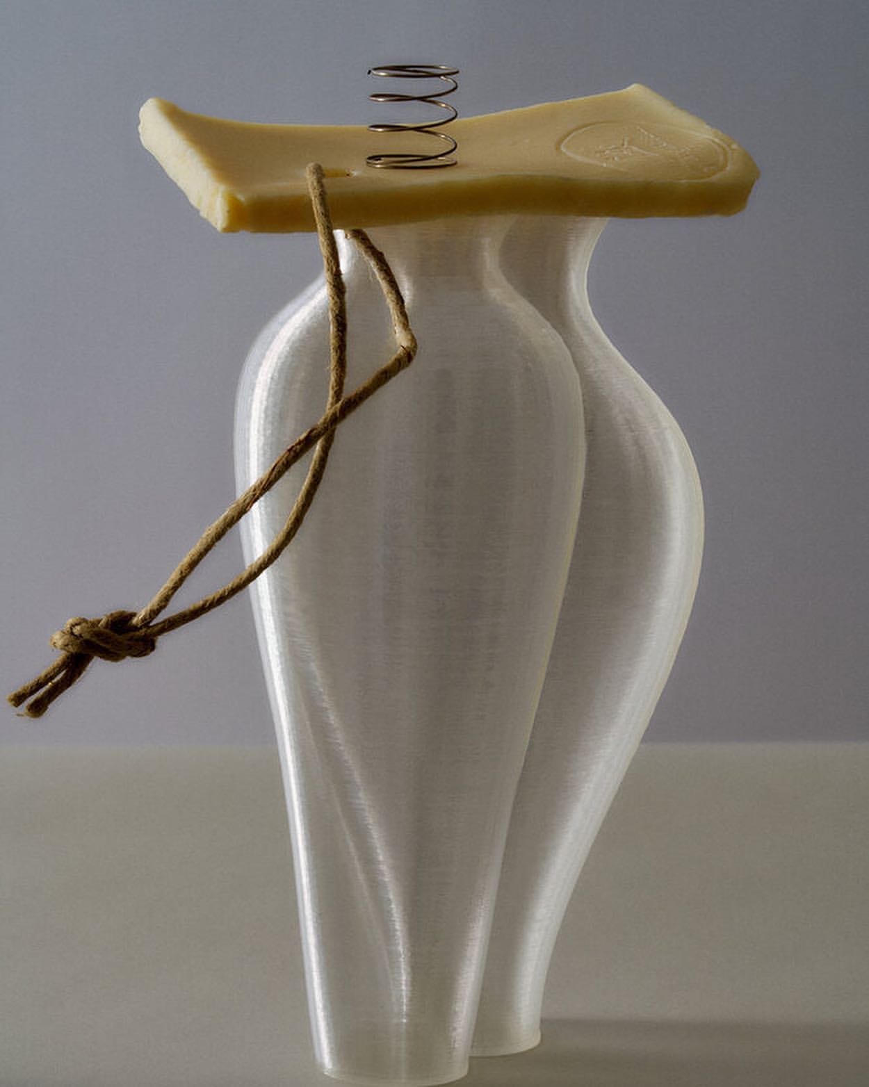 Still life of a white vase-like object with a piece of white cheese on top, supported by a spring, and tied with a brown string.