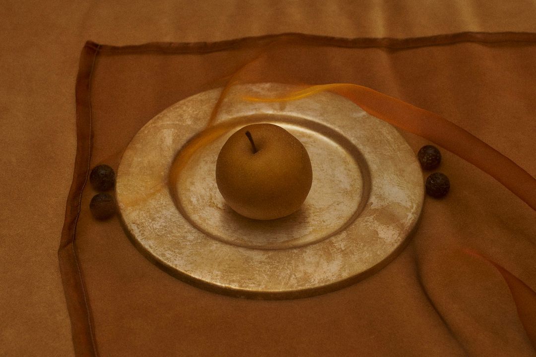 Golden Asian pear rests on a metallic gold plate alongside dark berries, set against warm brown fabric, captured in a still life shot.