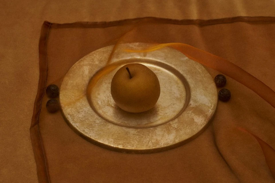 Golden Asian pear rests on a metallic gold plate alongside dark berries, set against warm brown fabric, captured in a still life shot.