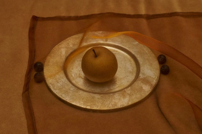 Golden Asian pear rests on a metallic gold plate alongside dark berries, set against warm brown fabric, captured in a still life shot.