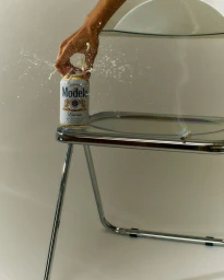 Hand splashing liquid onto a Modelo beer can resting on a chrome glass table by Heami Lee
