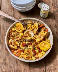 Ceramic bowl of pasta with clams, shrimp, corn, and lemon halves on a rustic wooden table, photographed by Heami Lee.