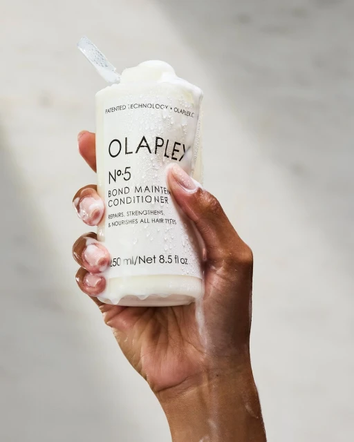 Hand holds Olaplex No. 5 Bond Maintenance Conditioner bottle with white lotion. Beauty product advert on light gray background.
