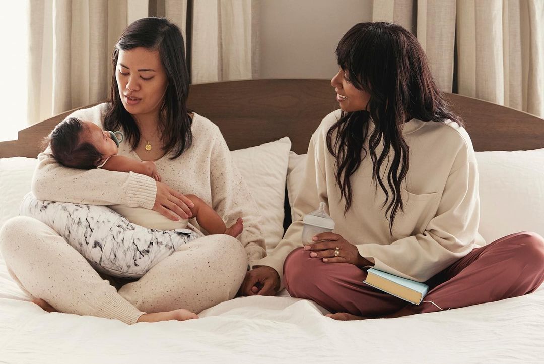 Two women sit cross-legged on a bed with a newborn baby and a baby bottle. Soft, neutral tones create a cozy, familial atmosphere.