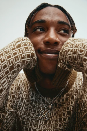 Close-up portrait of a Black man with braided hair wearing a patterned brown sweater and silver chain necklace against a light background.
