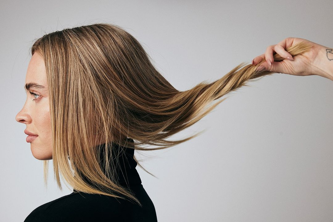 Profile of blonde woman in black top with another hand holding strand of her hair against grey backdrop in beauty shot.