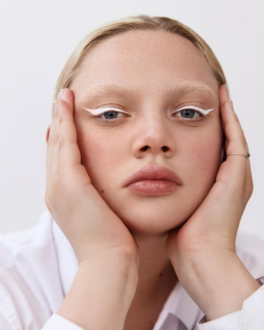 Close-up portrait of an adult woman with blonde hair and fair skin, wearing white eyeliner and a white shirt against a white background.