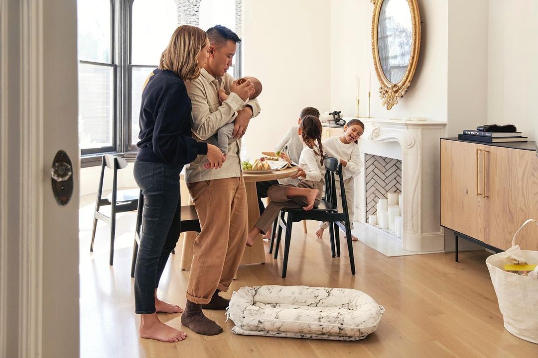 Family portrait indoors with two parents holding a newborn baby; three children in background, modern home interior, light wood floors.