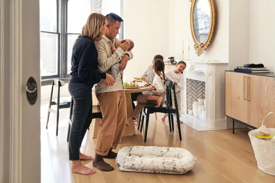 Family portrait indoors with two parents holding a newborn baby; three children in background, modern home interior, light wood floors.