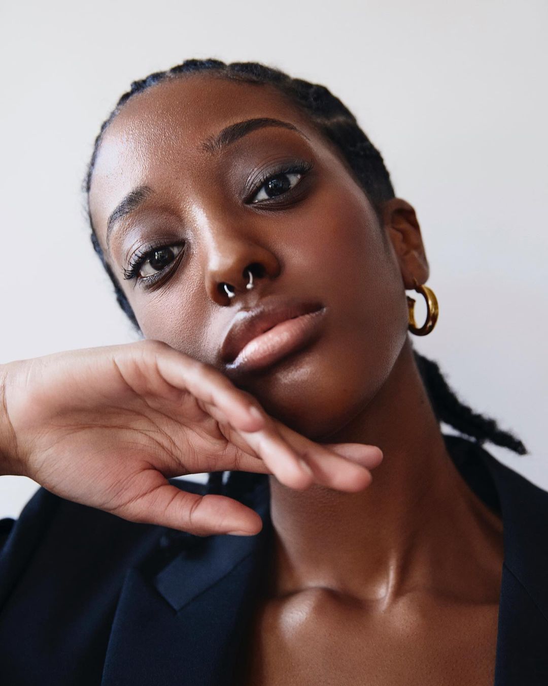Close-up portrait of a young Black woman with dark hair and a septum piercing, wearing a black jacket and gold hoop earrings.