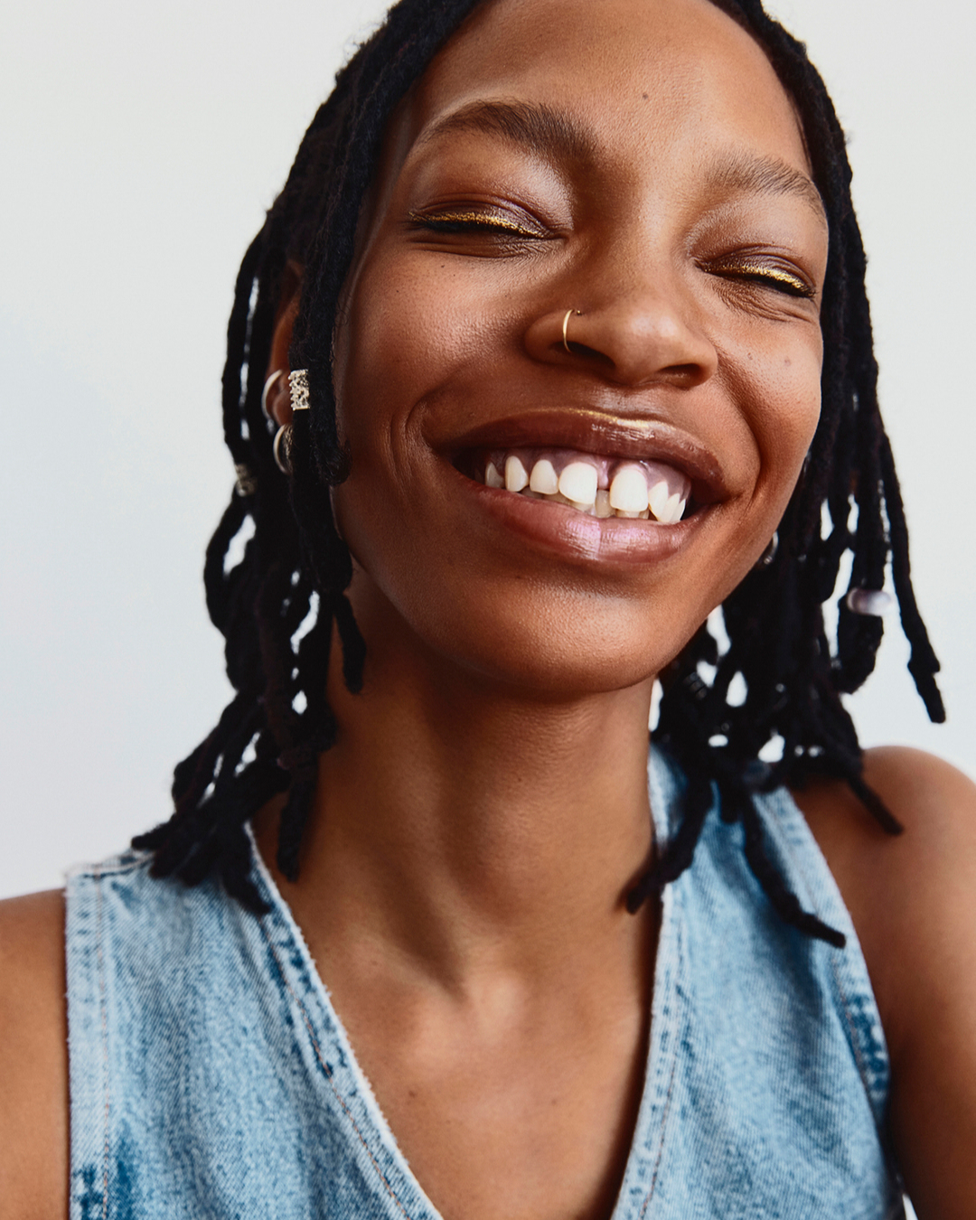 Close-up studio shot of a smiling adult female with dreadlocks wearing a denim top against a white backdrop.