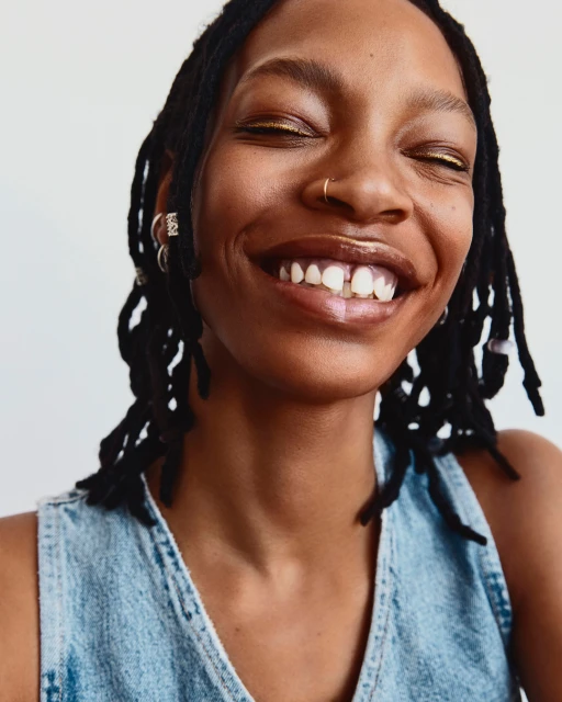 Close-up studio shot of a smiling adult female with dreadlocks wearing a denim top against a white backdrop.