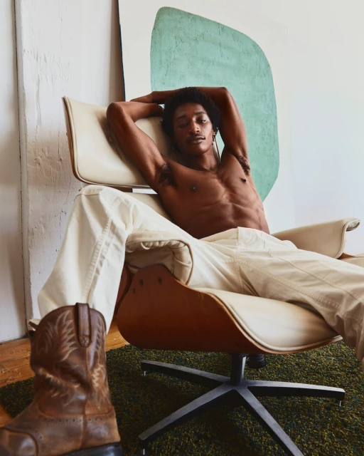 Man lounges shirtless in a modern chair with cowboy boots in the foreground. An abstract painting adds to the contemporary indoor scene.