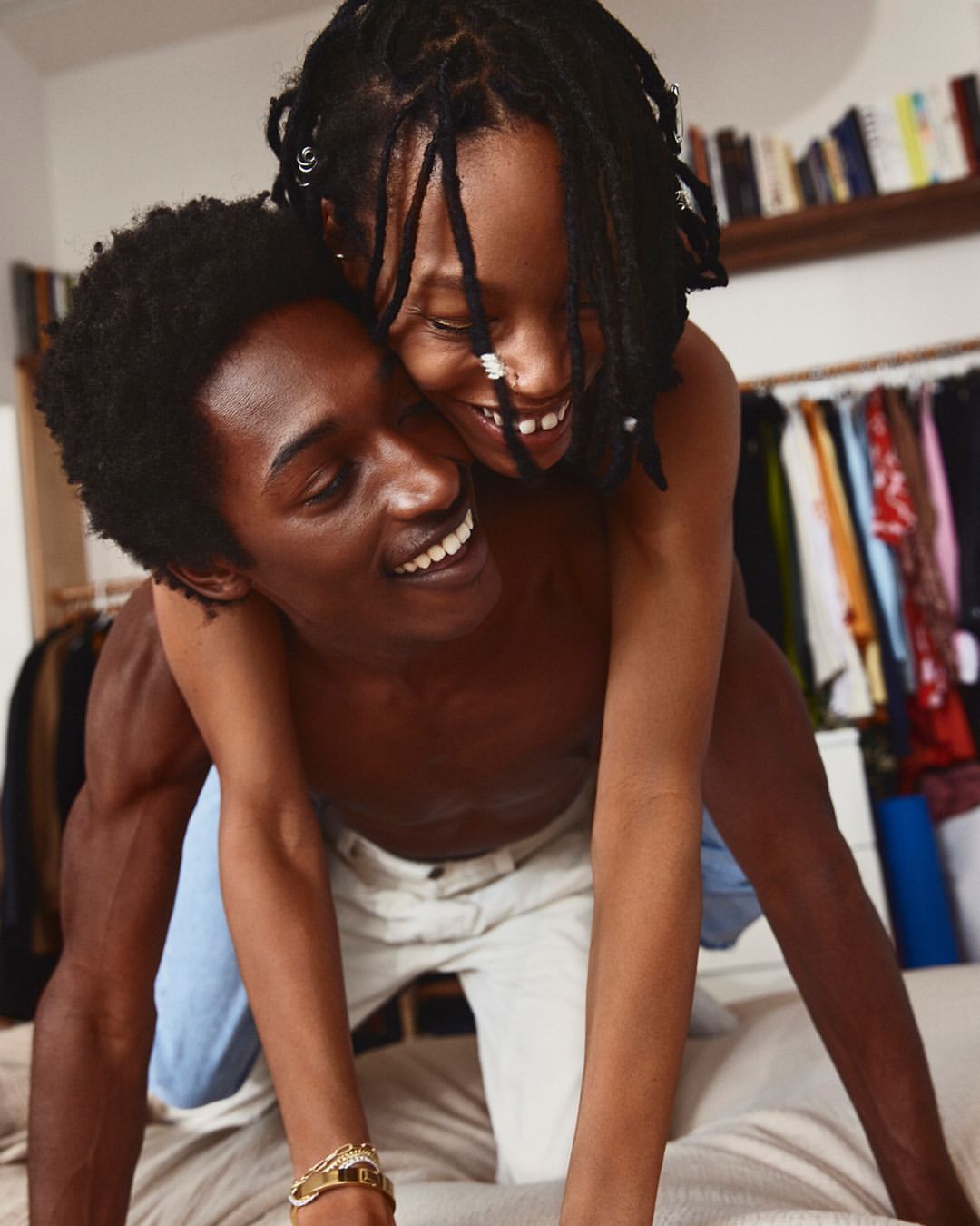 An African-American couple embraces indoors. The shirtless man kneels as his partner playfully jumps on his back, both smiling.
