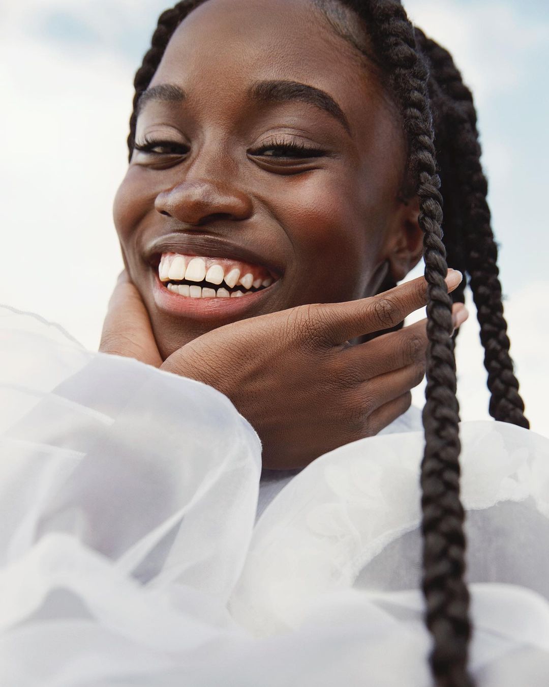 Close-up portrait of a smiling African-American woman with braids in a white flowing dress, set against a bright, airy sky background.