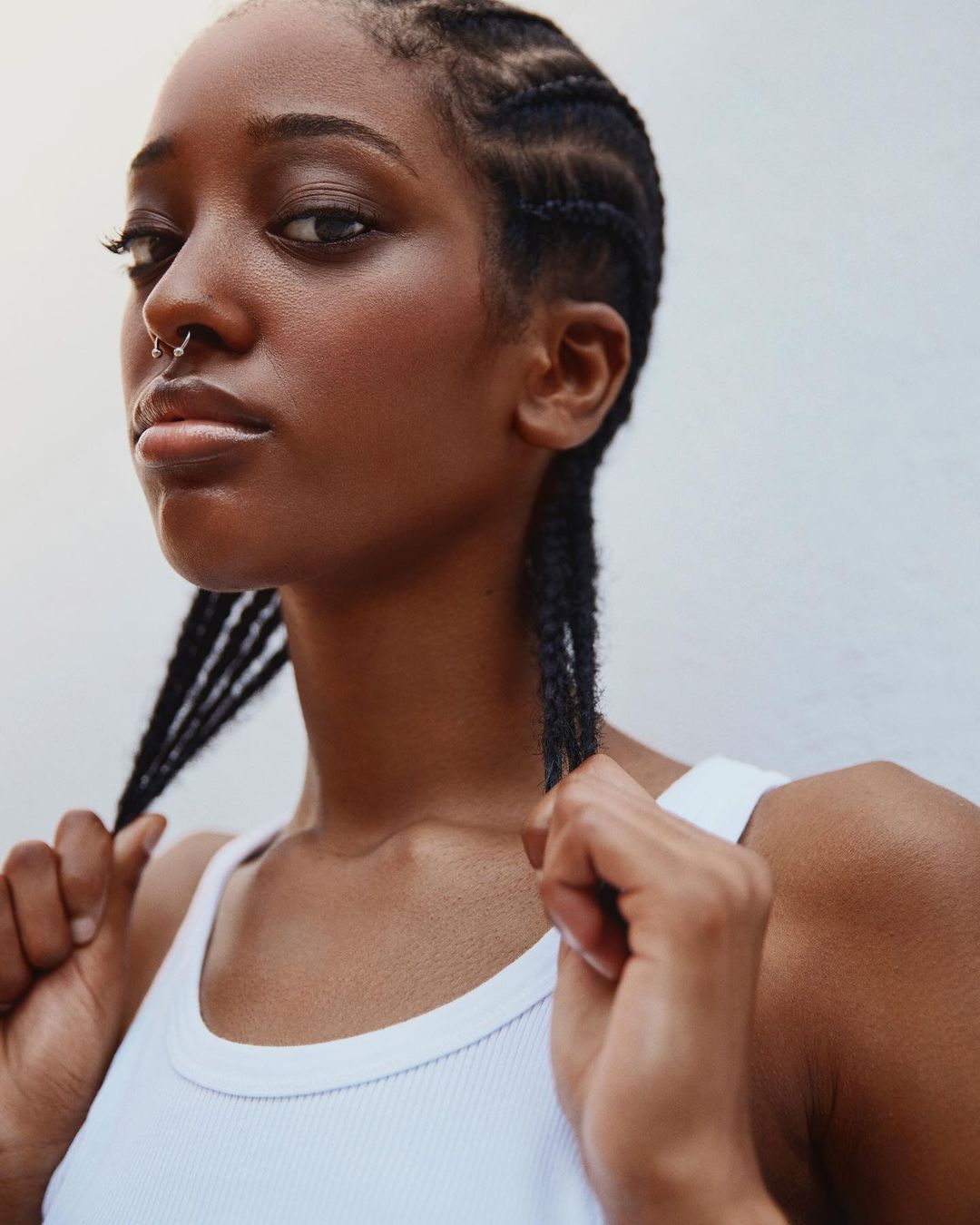 Portrait of a young African-American woman with braided hair and a nose ring, wearing a white ribbed tank top against a plain background.