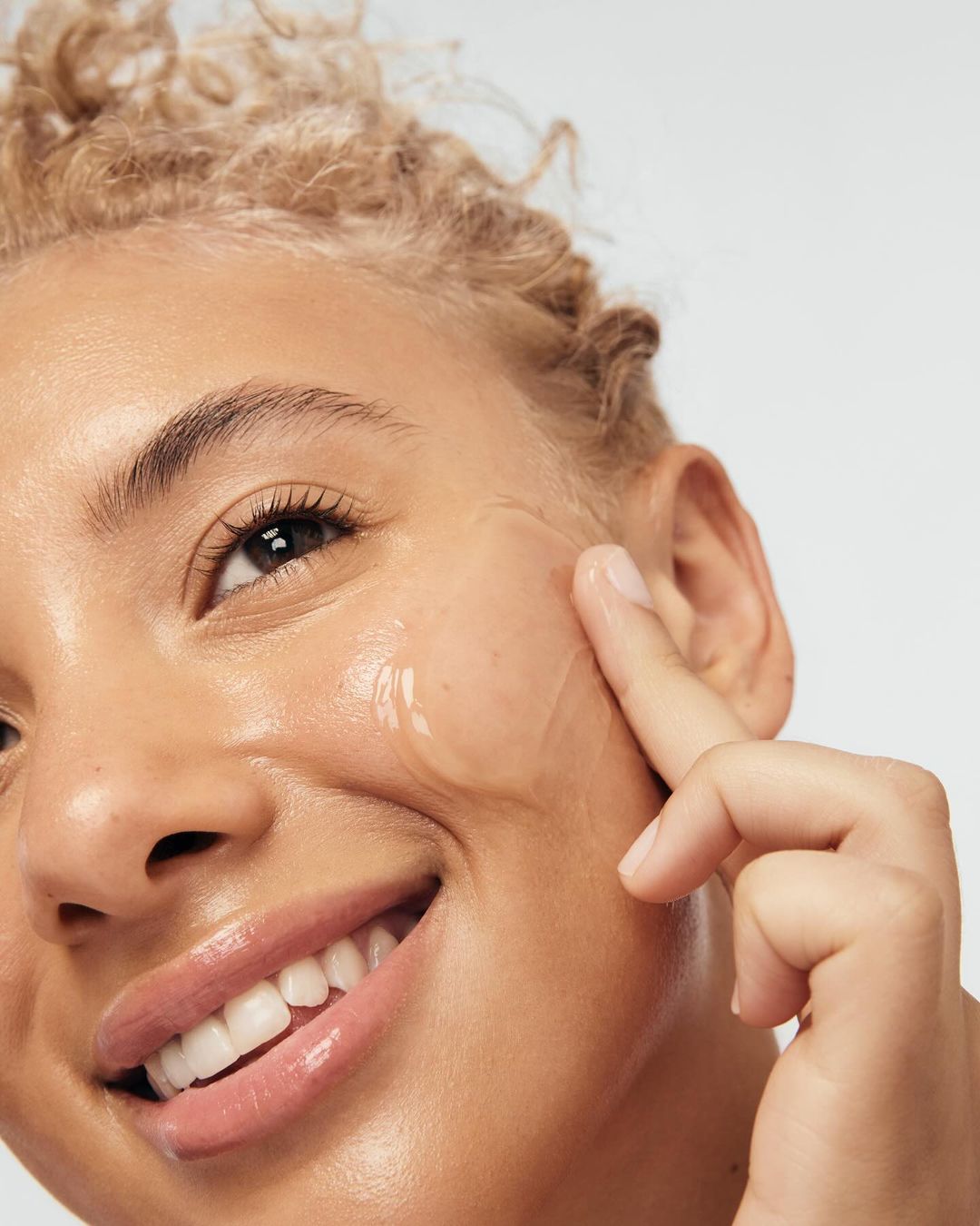 Close-up of a smiling adult woman with light skin, applying serum to her face against a light grey background, beauty skincare concept.