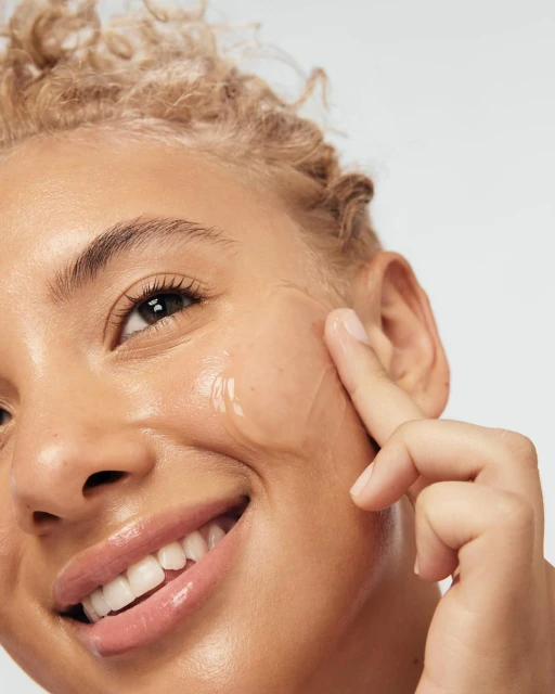 Close-up of a smiling adult woman with light skin, applying serum to her face against a light grey background, beauty skincare concept.