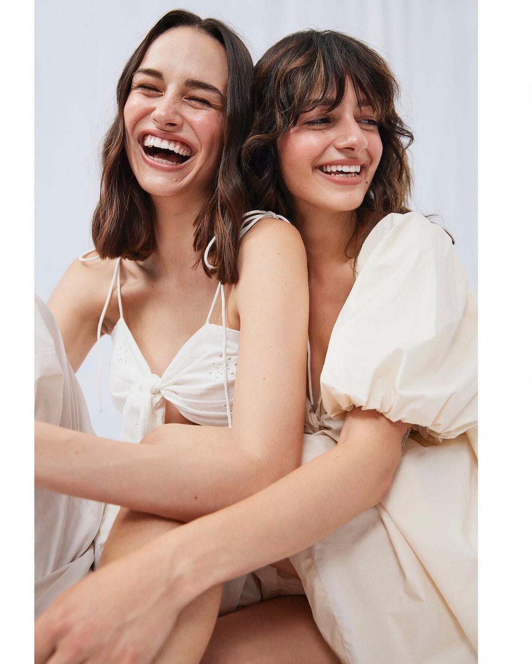 Two young women smile in an indoor studio shot. One wears a white eyelet crop top and pants, and the other wears a cream puff-sleeve dress.