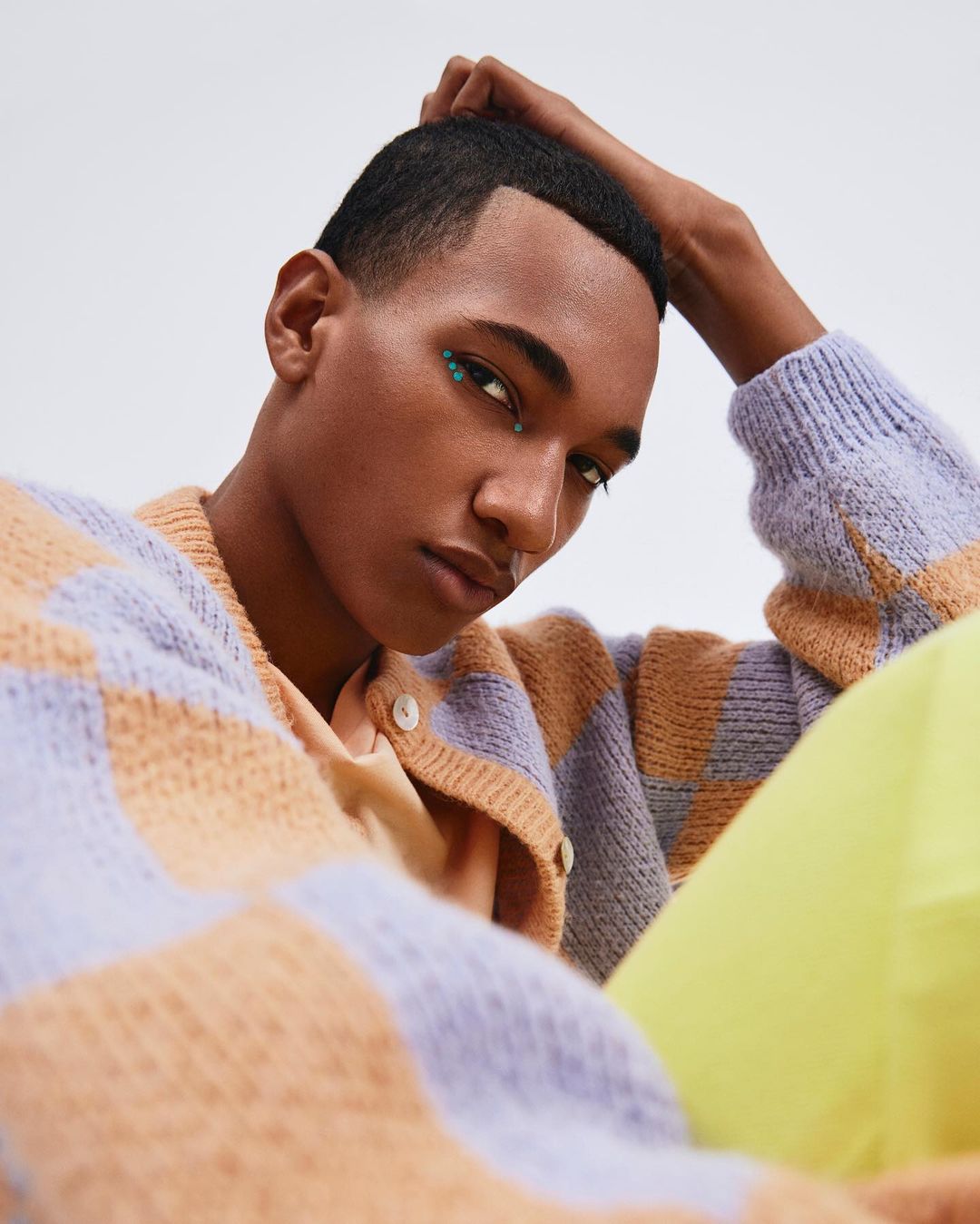 Portrait of an adult male with glitter eyeliner in a photo studio, wearing a patterned cardigan, neon yellow pants, against a white backdrop.
