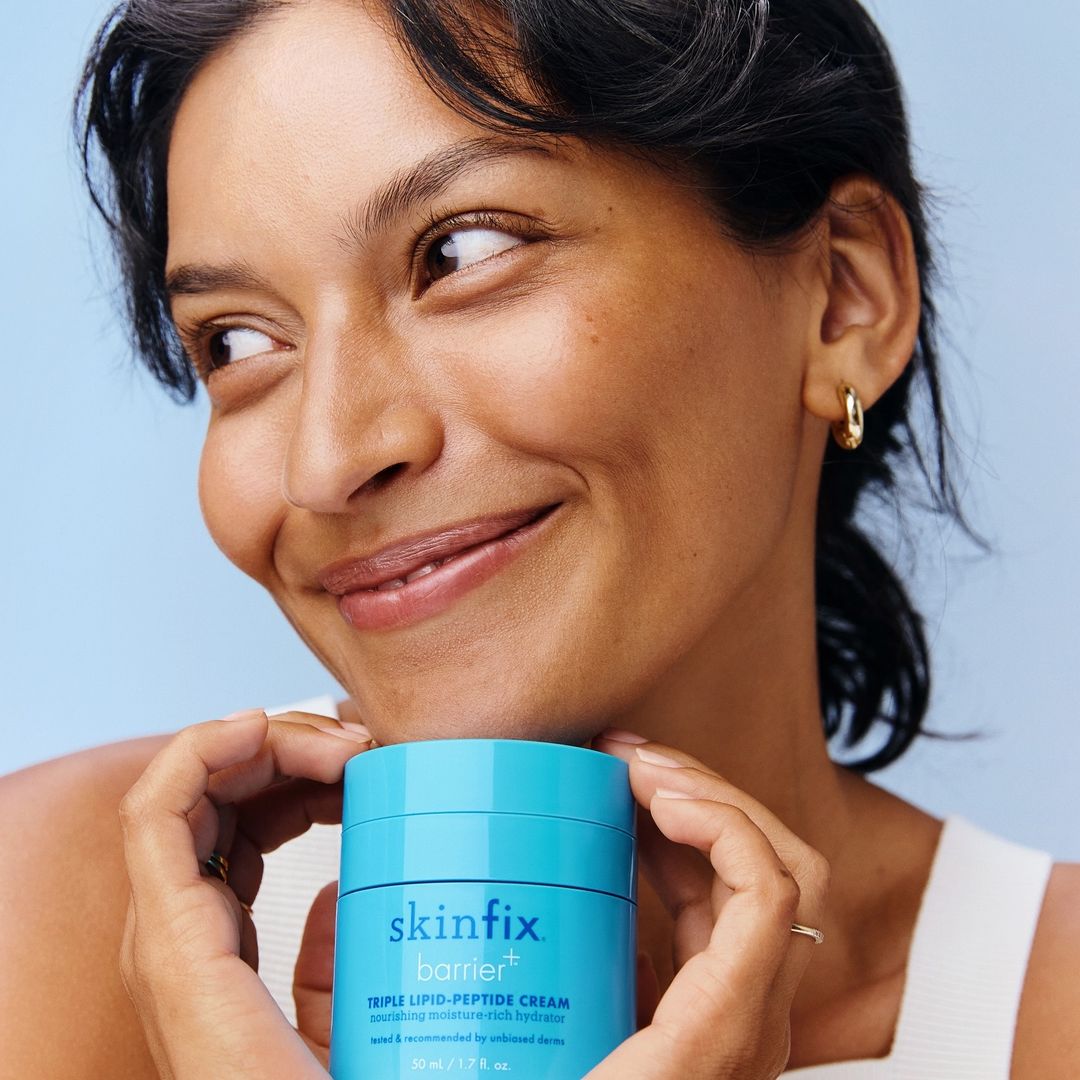 Close-up portrait of a smiling woman holding a blue jar of Skinfix barrier cream against a soft blue background.