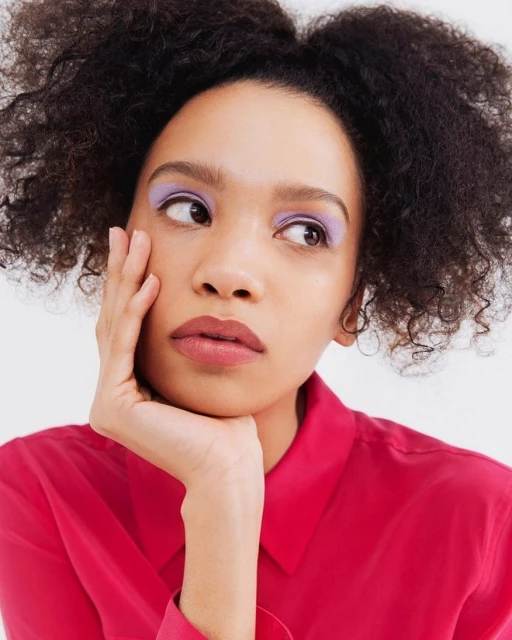 Portrait of a young adult female with an afro hairstyle wearing a pink button-down shirt and lavender eyeshadow, gazing to the side on a white background.