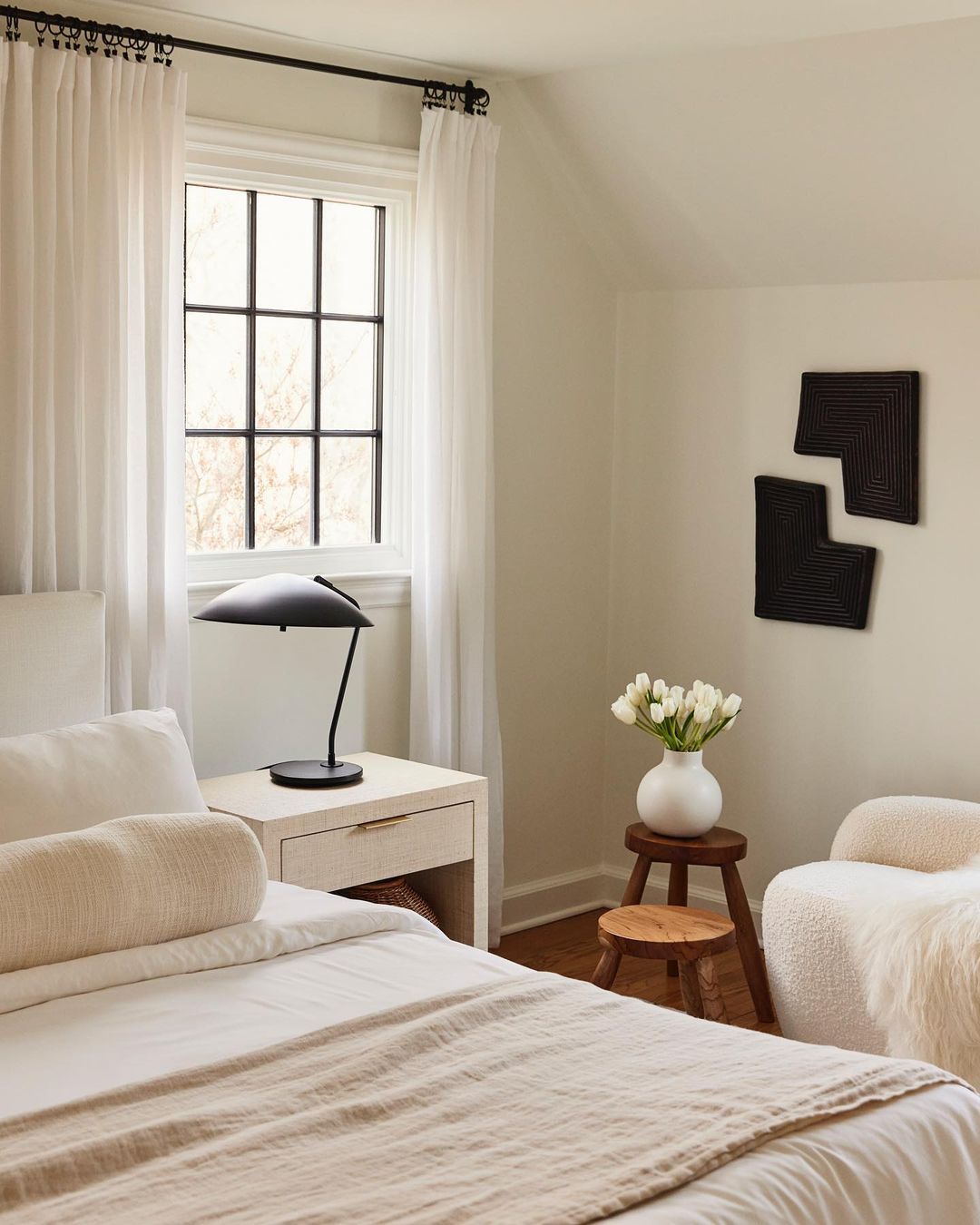 Bright, neutral bedroom with a white bed, a black lamp on a nightstand, a wooden stool with tulips, and abstract art on the wall.