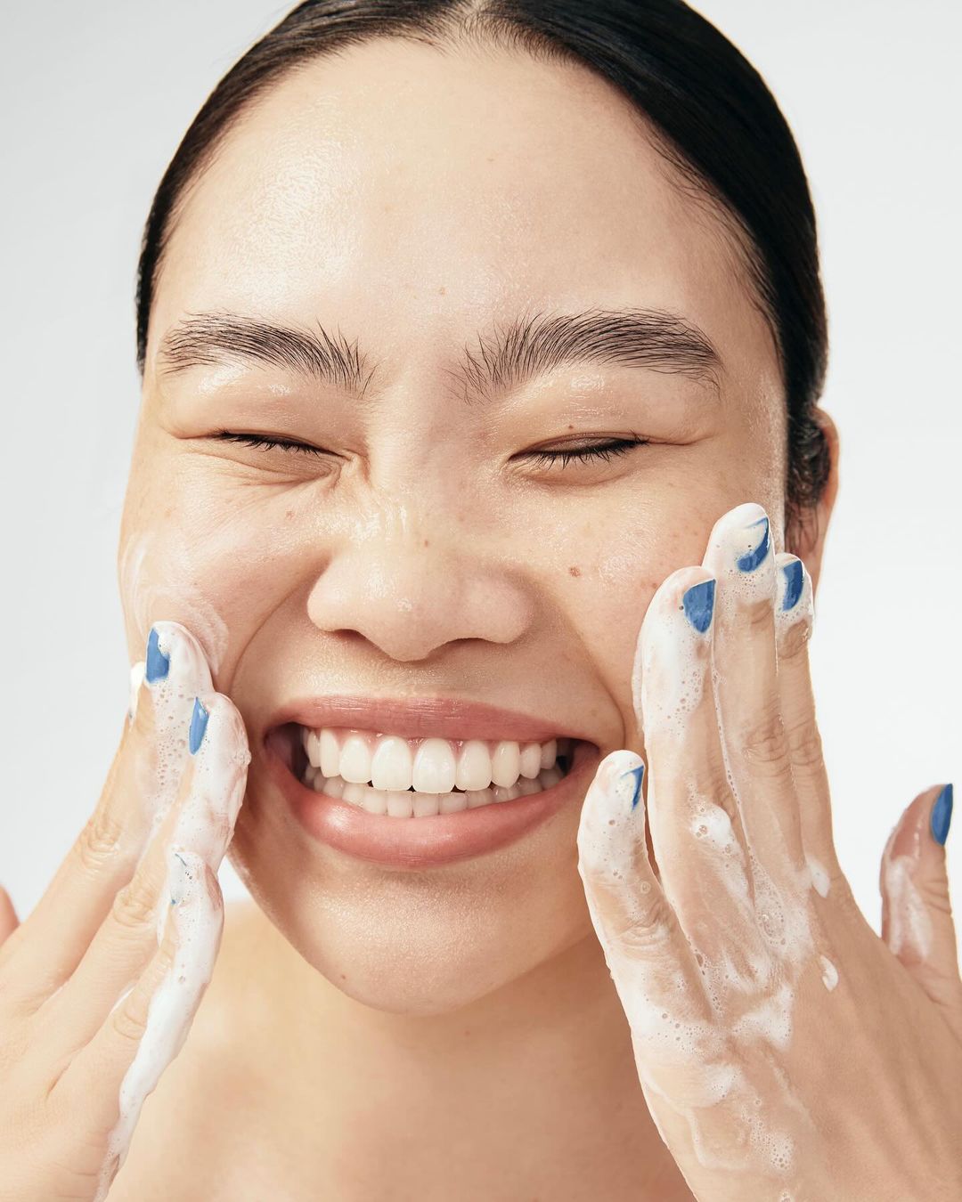Close-up of a smiling Asian woman with closed eyes, applying white foam cleanser to her face with both hands against a plain background.