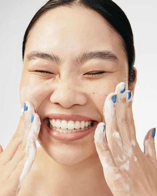Close-up of a smiling Asian woman with closed eyes, applying white foam cleanser to her face with both hands against a plain background.