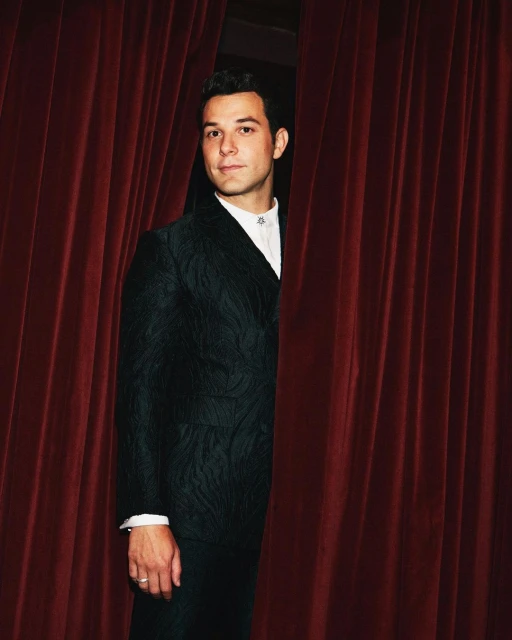 Headshot of a man wearing a floral-patterned suit posing next to red velvet curtains in a portrait, showcasing a formal and theatrical aesthetic.