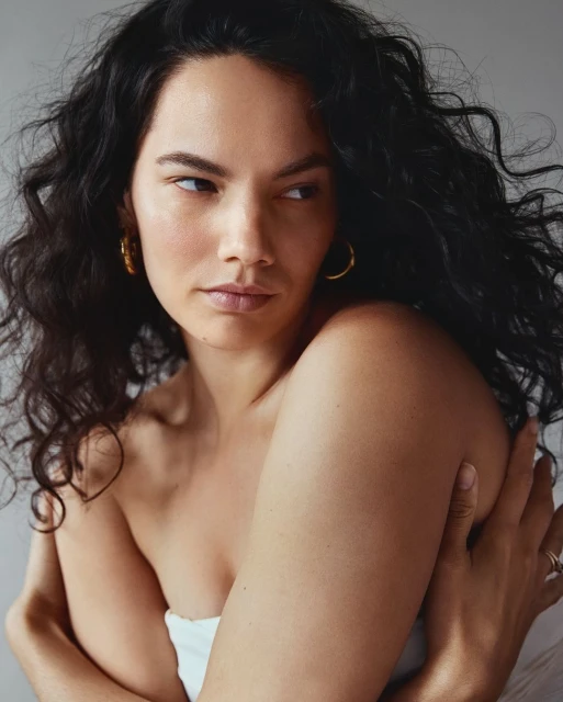 Portrait of a woman with dark curly hair and golden earrings, posing against a neutral background. She is wrapped in white fabric.