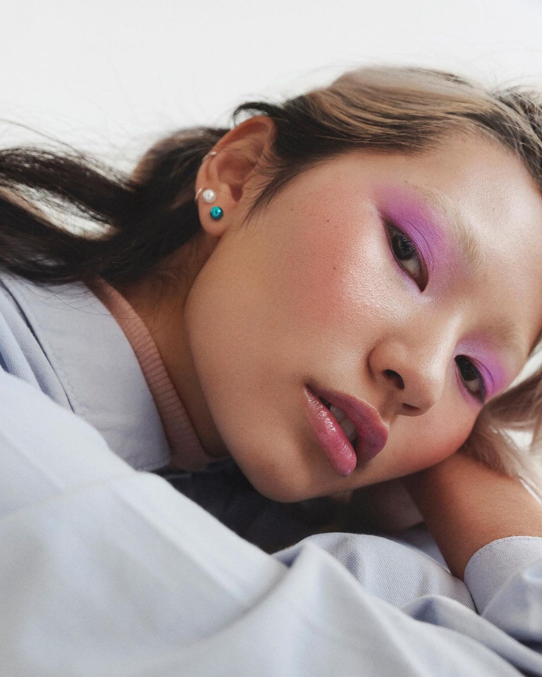Close-up of an Asian adult woman with vibrant purple eyeshadow and a light blue shirt, captured in soft, diffused lighting.