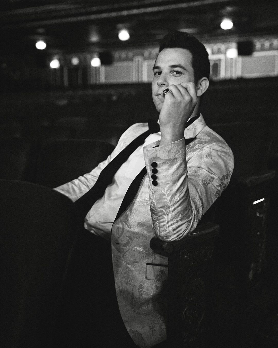 Black-and-white portrait of a man in a paisley jacket and bow tie, seated in an ornate theatre with decorative lighting and dark seating.