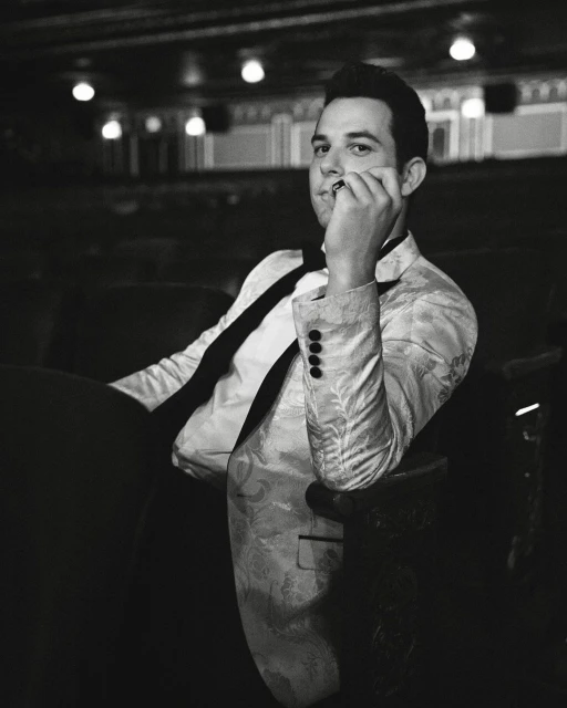 Black-and-white portrait of a man in a paisley jacket and bow tie, seated in an ornate theatre with decorative lighting and dark seating.
