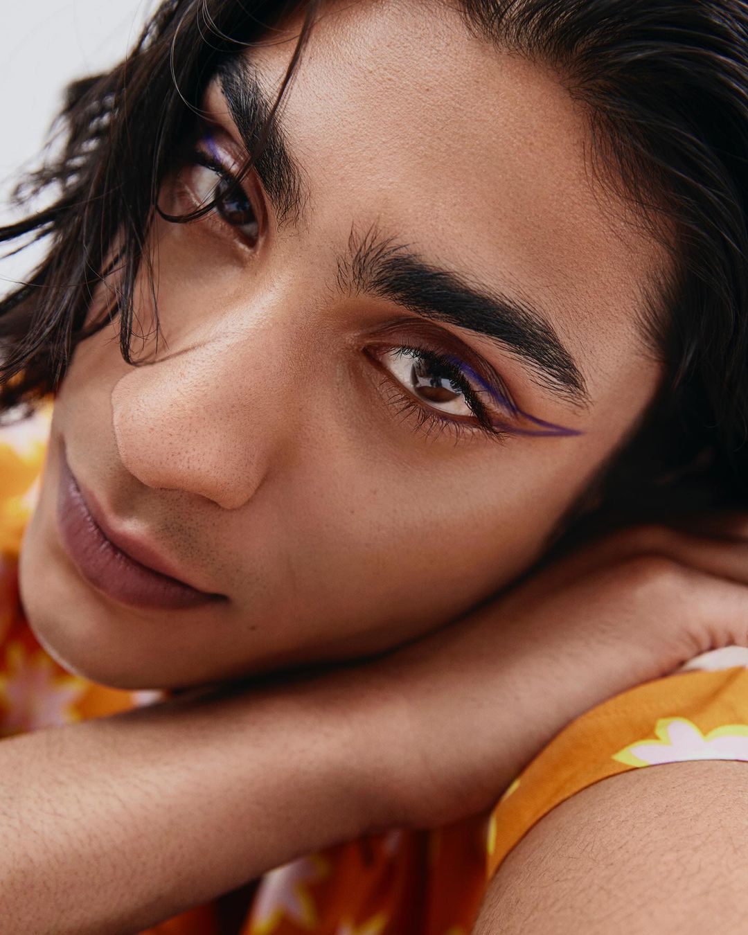 Close-up of a young adult with dark hair and purple eyeliner, resting their face on their arms wearing a floral-patterned shirt.