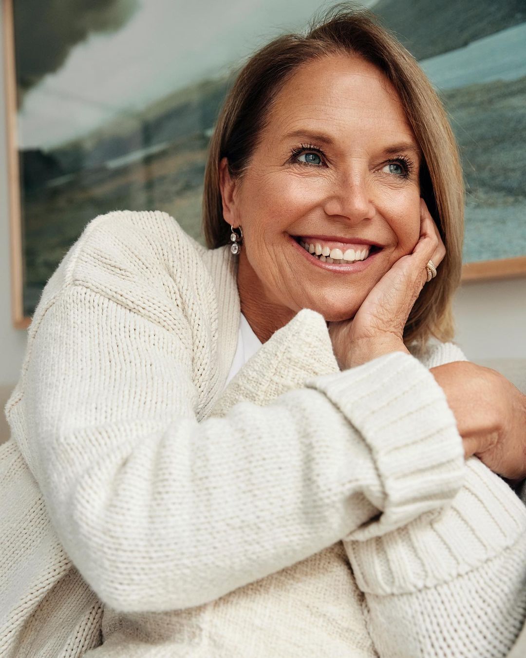 Portrait of an adult woman with short blonde hair, wearing a cream-colored sweater, smiling indoors near a decorative painting.