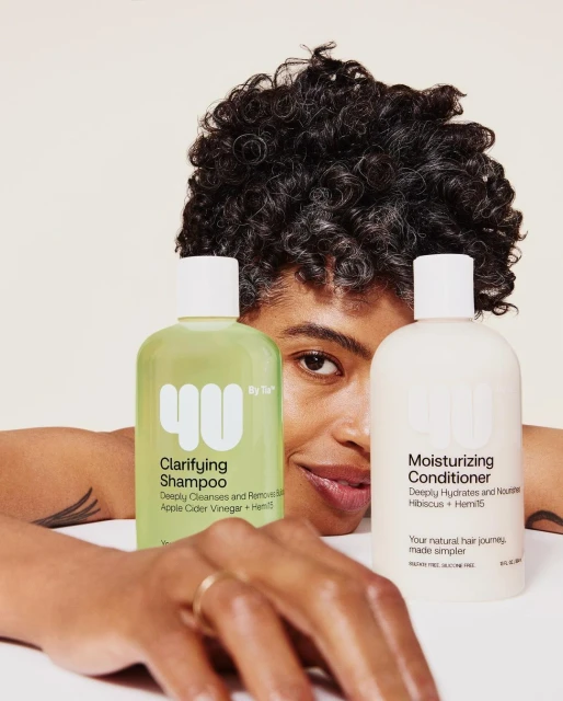 Person with curly hair smiles behind two '4U by Tia' hair product bottles, one green Clarifying Shampoo and one white Moisturizing Conditioner.