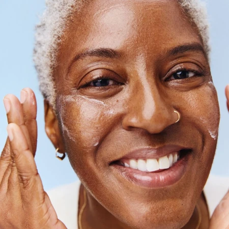 Close-up of a smiling, middle-aged woman with gray hair applying moisturizer to her face against a gradient light blue background.