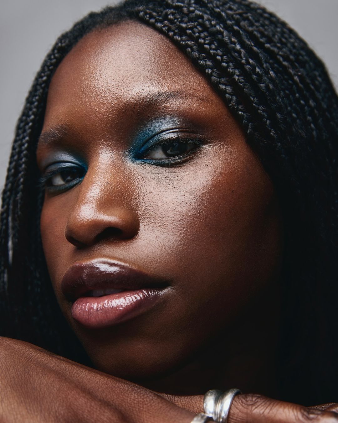 Close-up portrait of an adult woman with dark skin and braided hair, accented by teal eyeshadow and glossy lips, gazing directly at the viewer.