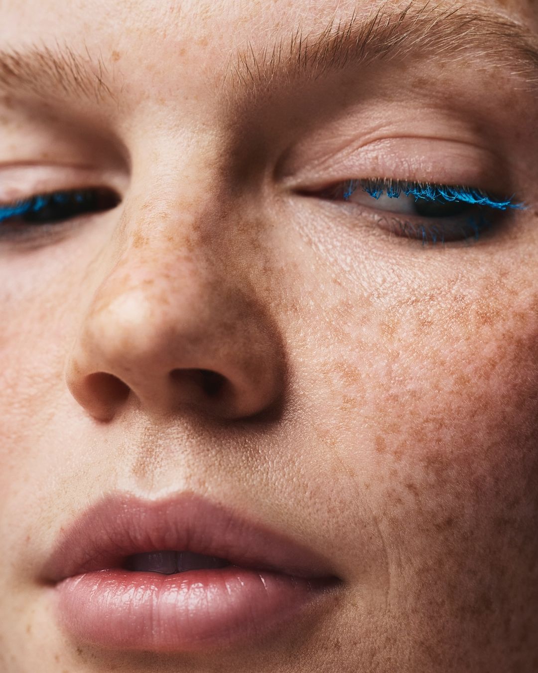 Close-up portrait of a young adult female with freckles and blue mascara on her eyelashes, focusing on beauty and personal care.