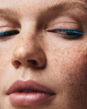 Close-up portrait of a young adult female with freckles and blue mascara on her eyelashes, focusing on beauty and personal care.